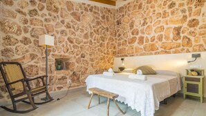 A bedroom with a stone wall, a bed with a white bedspread, a wooden rocking chair, a bedside table with a lamp, and a small cabinet.