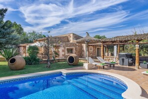 A swimming pool with a tiled edge, a stone building with a tiled roof, and outdoor seating with umbrellas.
