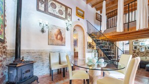 A cozy living room with a glass table, comfortable chairs, a vintage stove, and a staircase leading to an upper level. - Cas Mostatxet (sencelles) - Villa With Pool (Sencelles, Illes Balears)