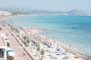 A beach with people sunbathing and swimming, a promenade with umbrellas, and buildings in the background. - Ca'n Pere Roc - Apartment In Can Picafort (Can Picafort, Illes Balears)