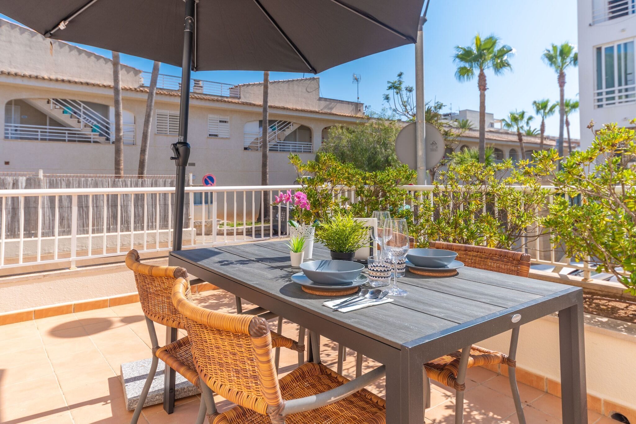 An outdoor patio with a table set for two, surrounded by wicker chairs, under a large umbrella.