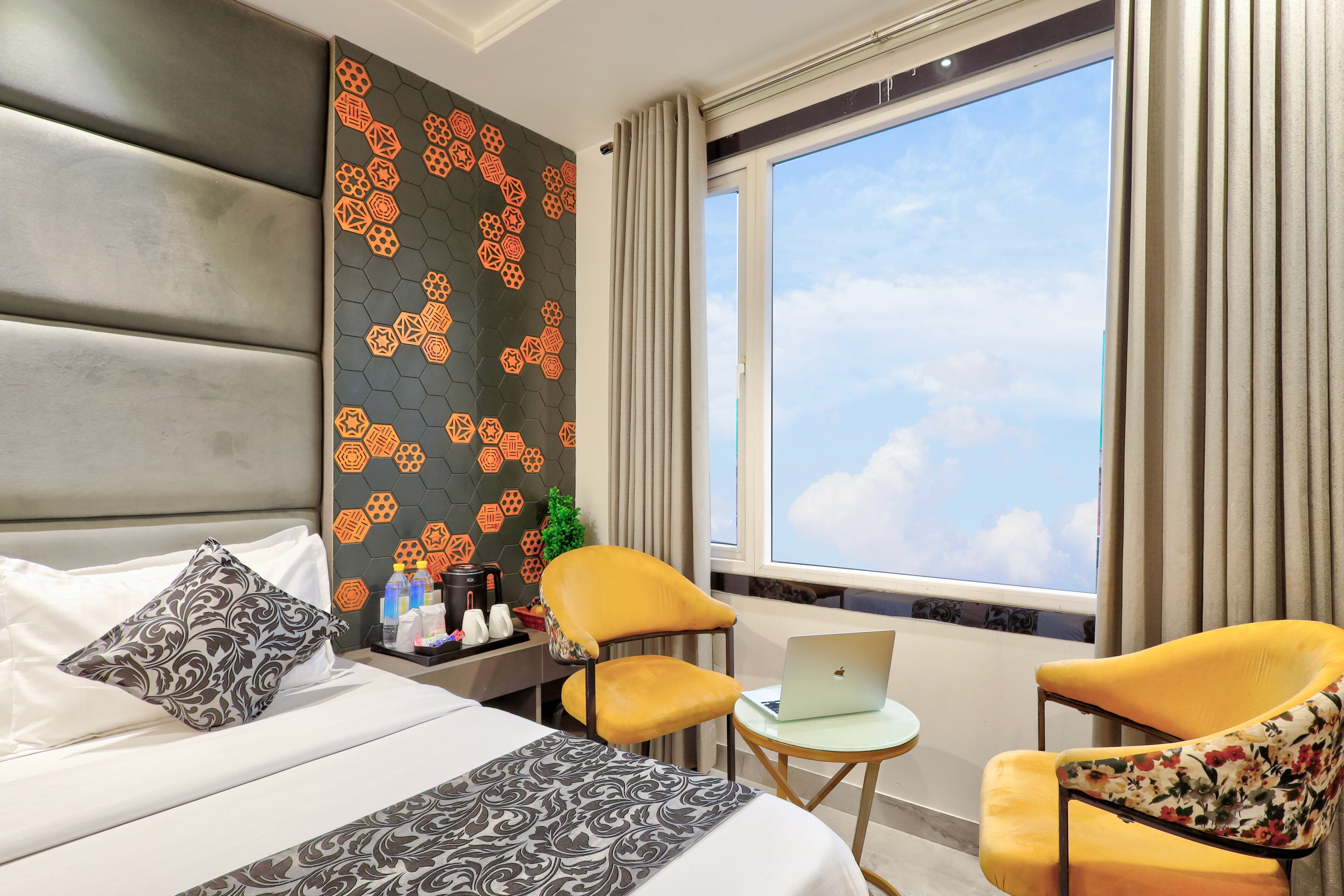 A hotel room with a bed, two chairs, a small table with a laptop, a decorative wall, and a large window with curtains.