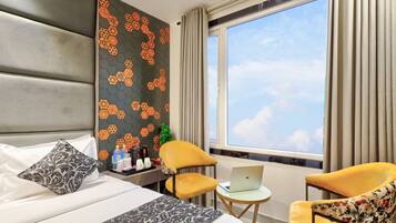 A hotel room with a bed, two chairs, a small table with a laptop, a decorative wall, and a large window with curtains.