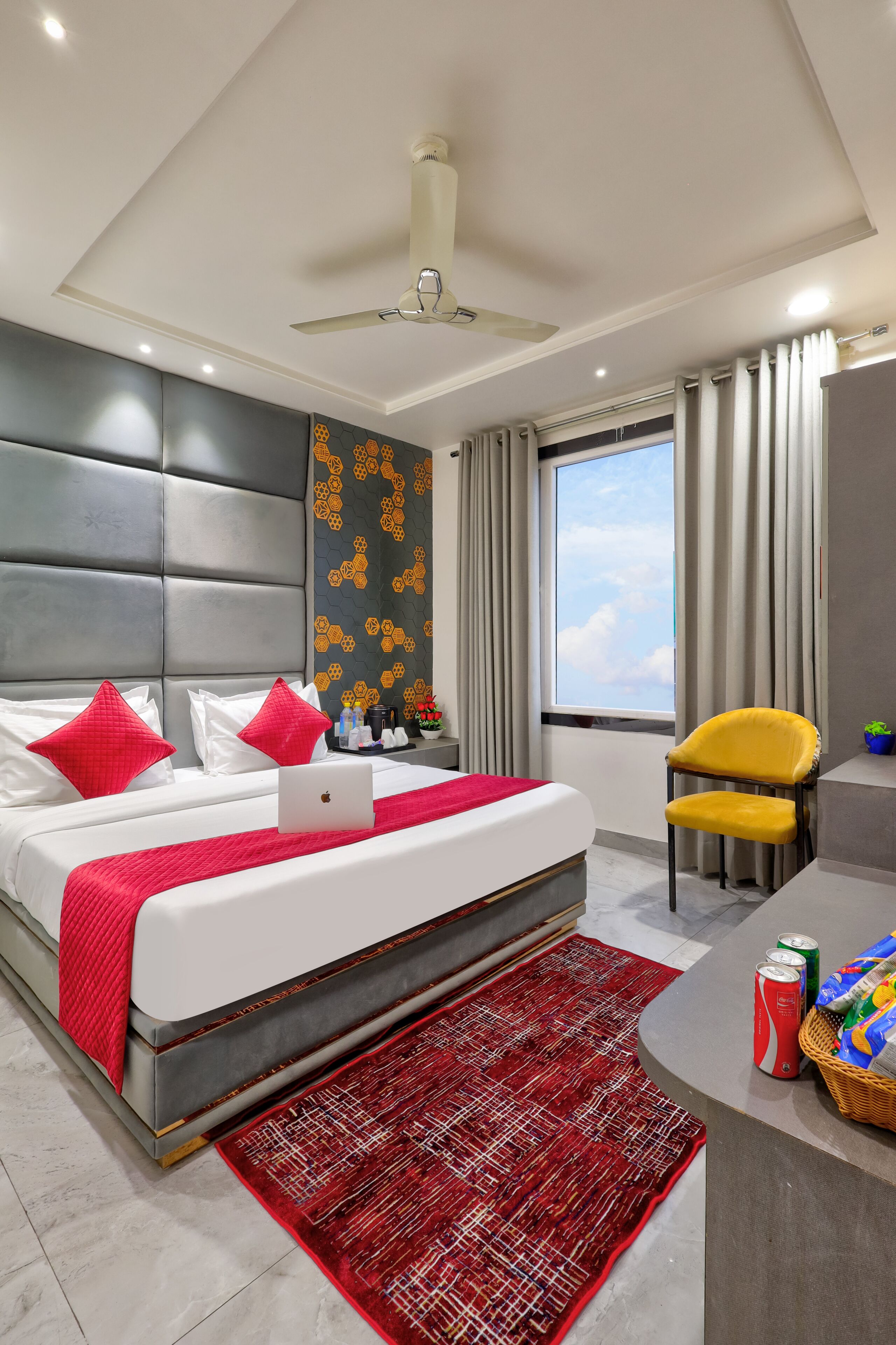 A modern hotel room with a bed, a desk, a chair, a laptop, a red rug, and a window with curtains.