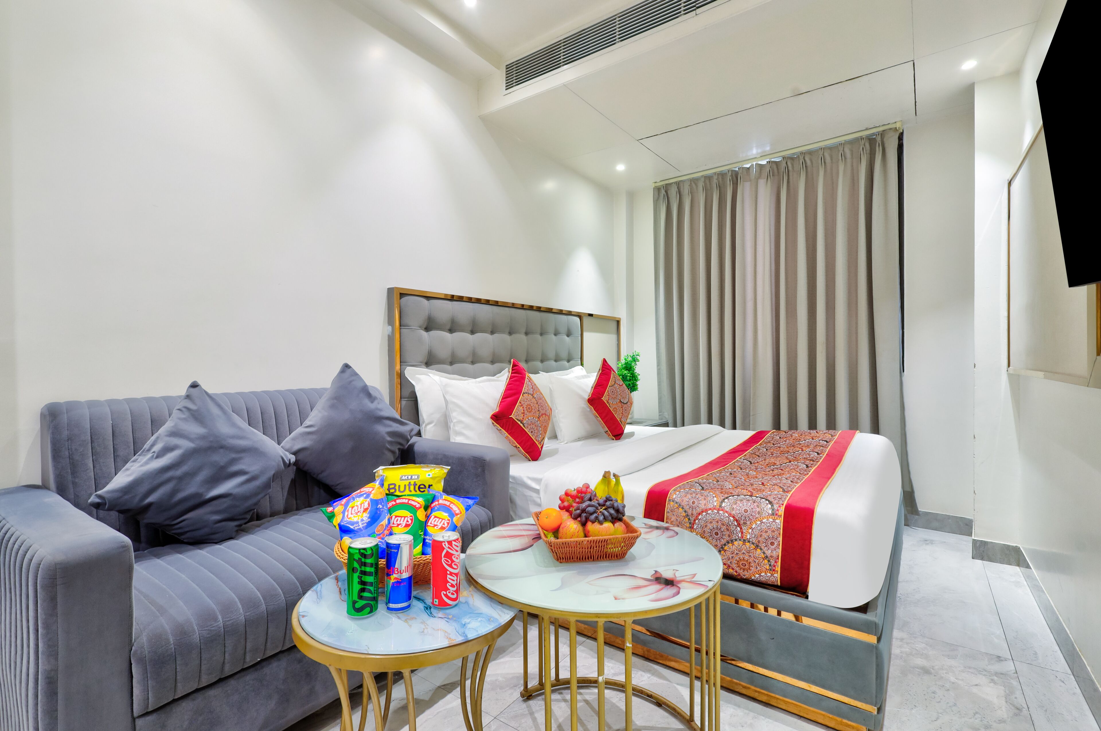 Quarto de hotel moderno com um sofá cinzento, uma cama com colcha estampada em vermelho e branco, uma mesa redonda pequena com lanches e uma janela grande com cortinas.