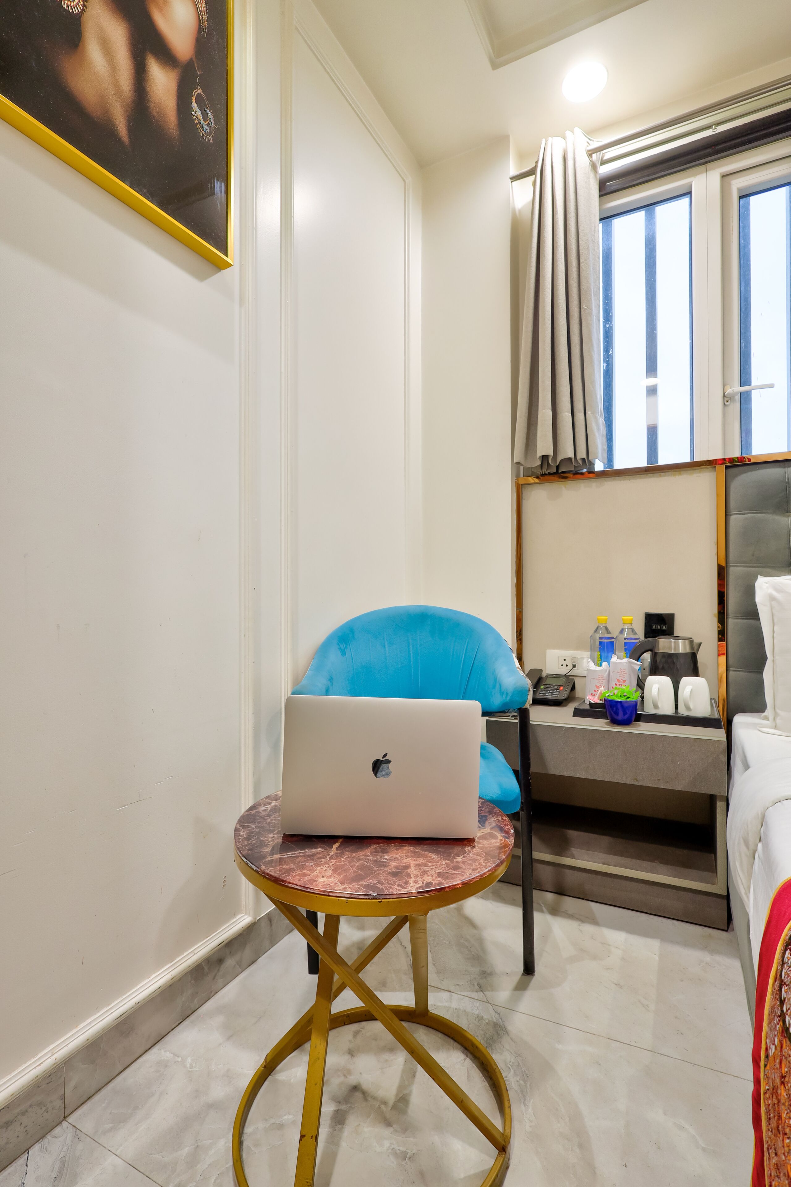 A room with a laptop on a small round table, a blue chair, a bed, a window with curtains, and a bedside table with toiletries.