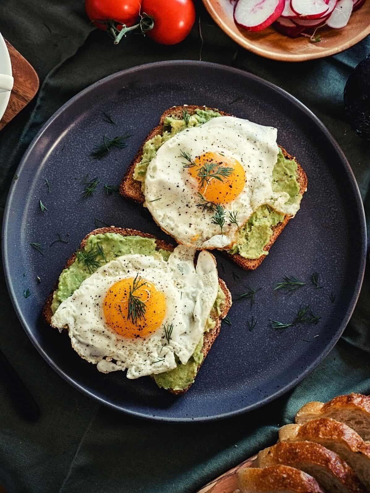 Zwei Toastscheiben mit Avocado und Spiegeleiern auf einem dunklen Teller.