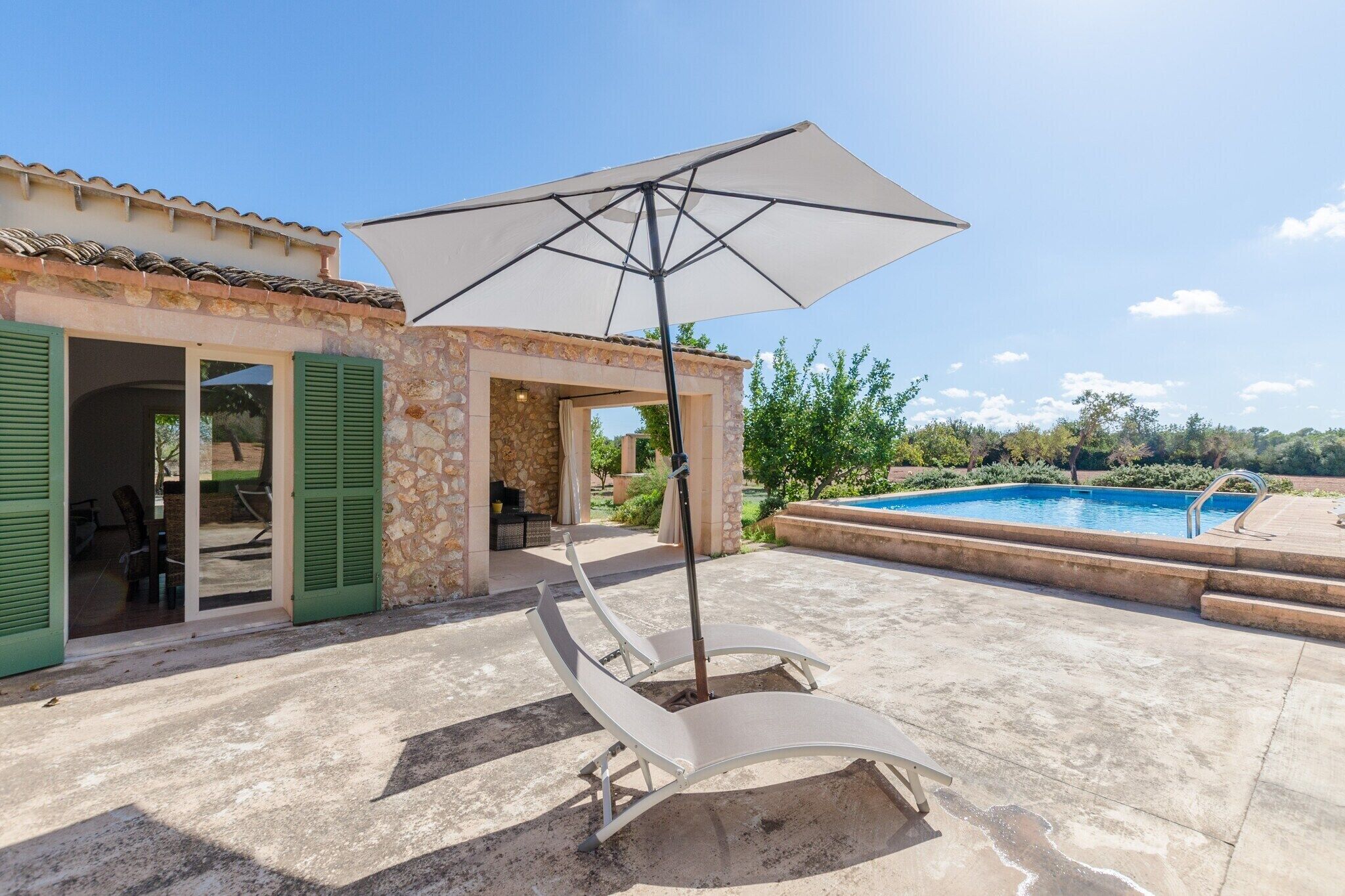 A poolside area with a lounge chair and umbrella, a stone building with green shutters, and a swimming pool.