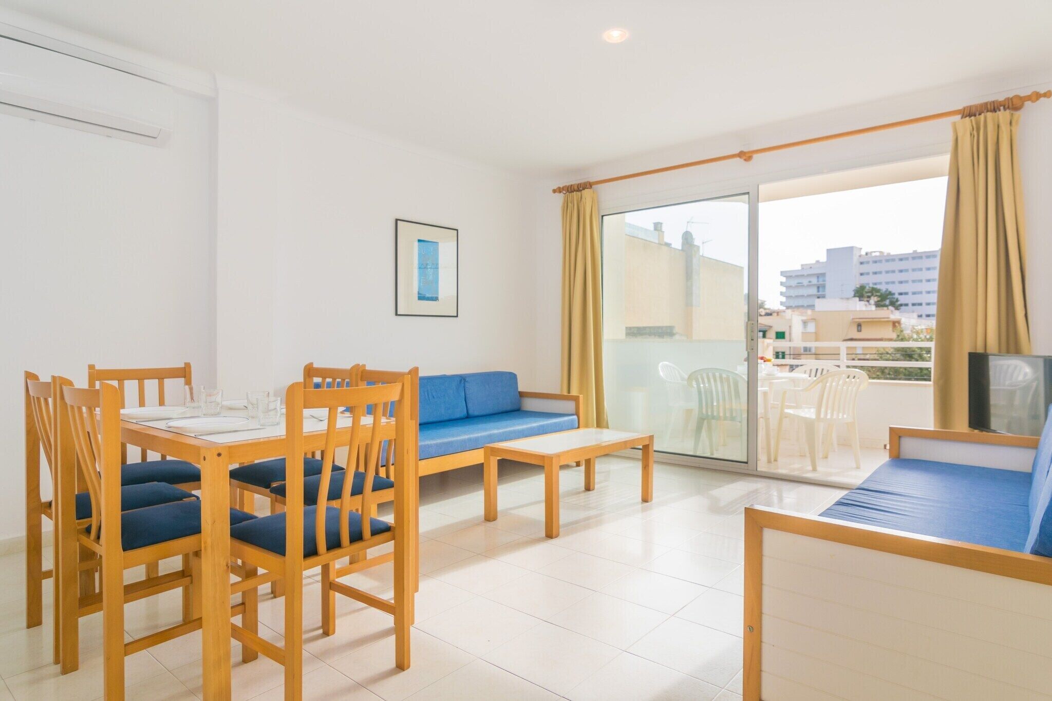 A room with a dining table, blue cushioned chairs, a sofa, a bed, and a balcony with a view of buildings.