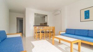 A modern living room with a blue sofa, a wooden dining table with chairs, and a kitchen visible through an open doorway. - Massanet (1c) - Apartment In Canyamel. Free Wifi (Canyamel, Illes Balears)
