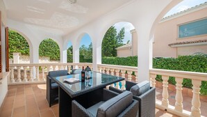 Un patio exterior cubierto con una mesa de vidrio y sillas de mimbre, rodeado de balaustradas blancas y follaje.
