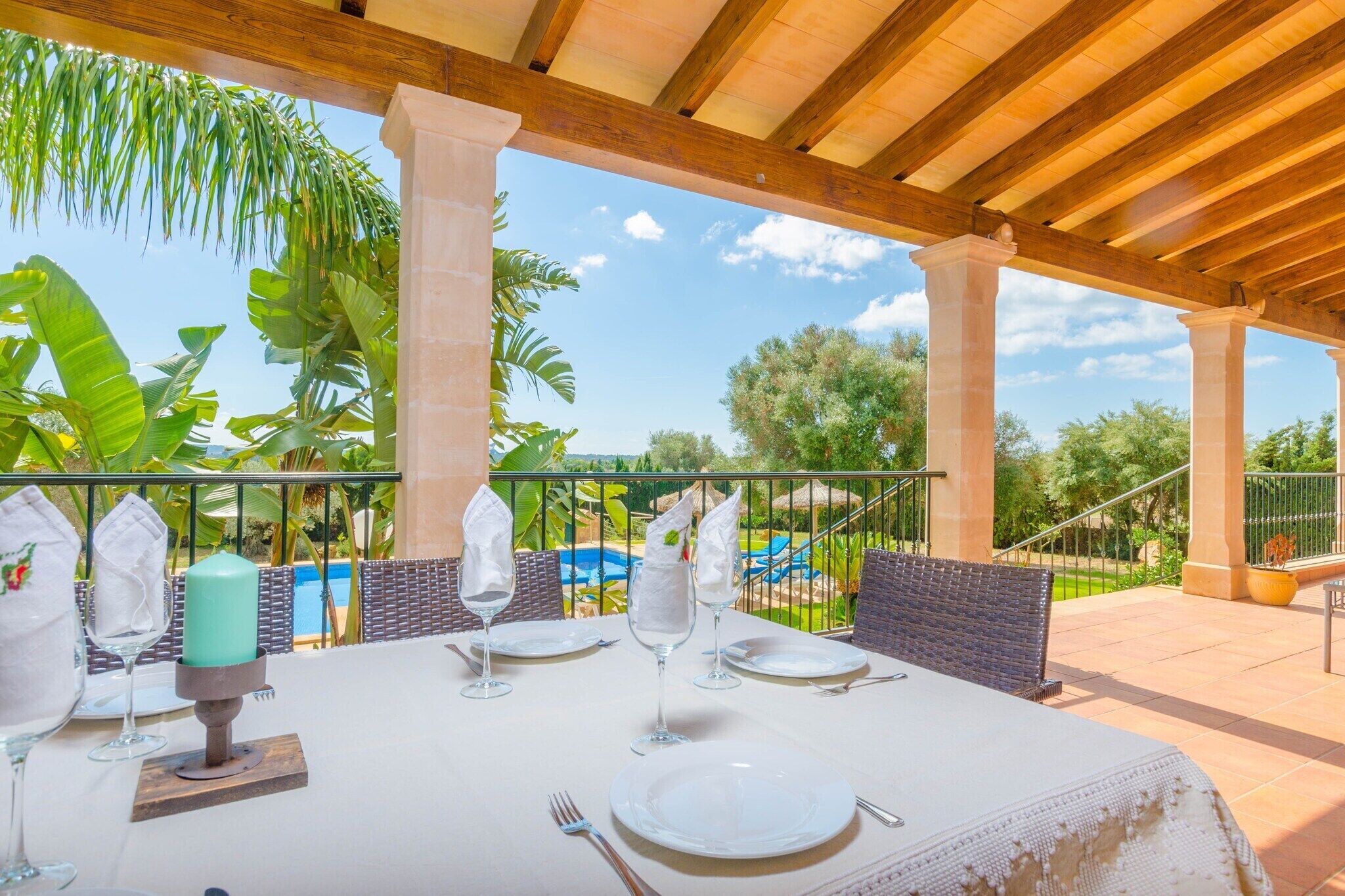 An outdoor dining area with a table set for four, surrounded by wicker chairs, under a wooden pergola with a view of a swimming pool and lush greenery.