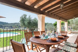 Eine Terrasse im Freien mit einem Holztisch, gedeckt für ein Essen, umgeben von Stühlen, mit Blick auf einen Swimmingpool und üppiges Grün.