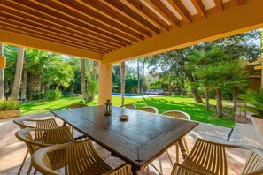 Patio esterno con tavolo e sedie in legno, circondato da vegetazione lussureggiante e una piscina.