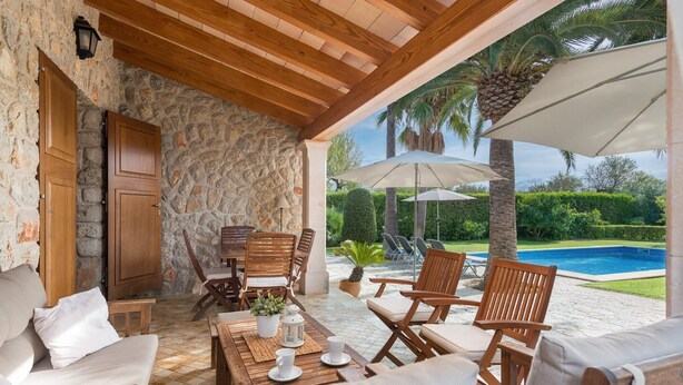An outdoor patio with wooden furniture, a stone wall, and a pool in the background. - Finca Pou Nou - Villa With Private Pool In Inca (Inca, Illes Balears)