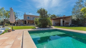 A swimming pool with a tiled edge, surrounded by a stone patio. There are outdoor umbrellas and a well-maintained lawn.