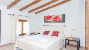 A bedroom with a large bed, wooden beams on the ceiling, a painting of red flowers, and a nightstand with a lamp.