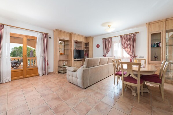 A living room with a sofa, dining table, chairs, and a TV. There is a sliding glass door leading to an outdoor area.