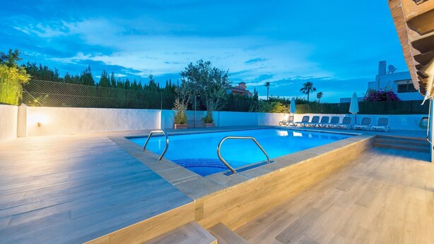 A swimming pool with a wooden deck, lounge chairs, and a view of a building in the background.