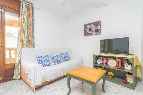 A living room with a sofa, a wooden table, a TV on a shelf, and a door with a curtain. - Casa Manolo - Apartment In Cala Lliteres (Cala Ratjada, Illes Balears)