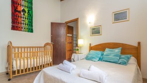 Ein Schlafzimmer mit einem Holz-Babybett, einem Bett mit weißer Bettwäsche und blauen Kissen, Wandteppich und einem kleinen Tisch mit Lampe.