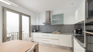 Cocina moderna con gabinetes blancos, extractor de acero inoxidable y horno empotrado. Hay una mesa de comedor con sillas y una ventana con vistas a un balcón.