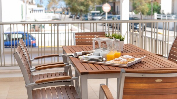 Una mesa de madera con alimentos para el desayuno, una planta en maceta y vista a la calle.