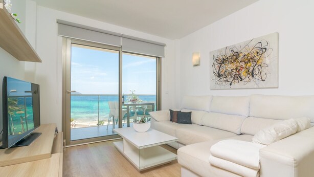 Ein modernes Wohnzimmer mit einem weißen Sofa, einem Couchtisch und einem Fernseher auf einem Holzboden. Ein großes Fenster bietet Blick auf Strand und Meer.