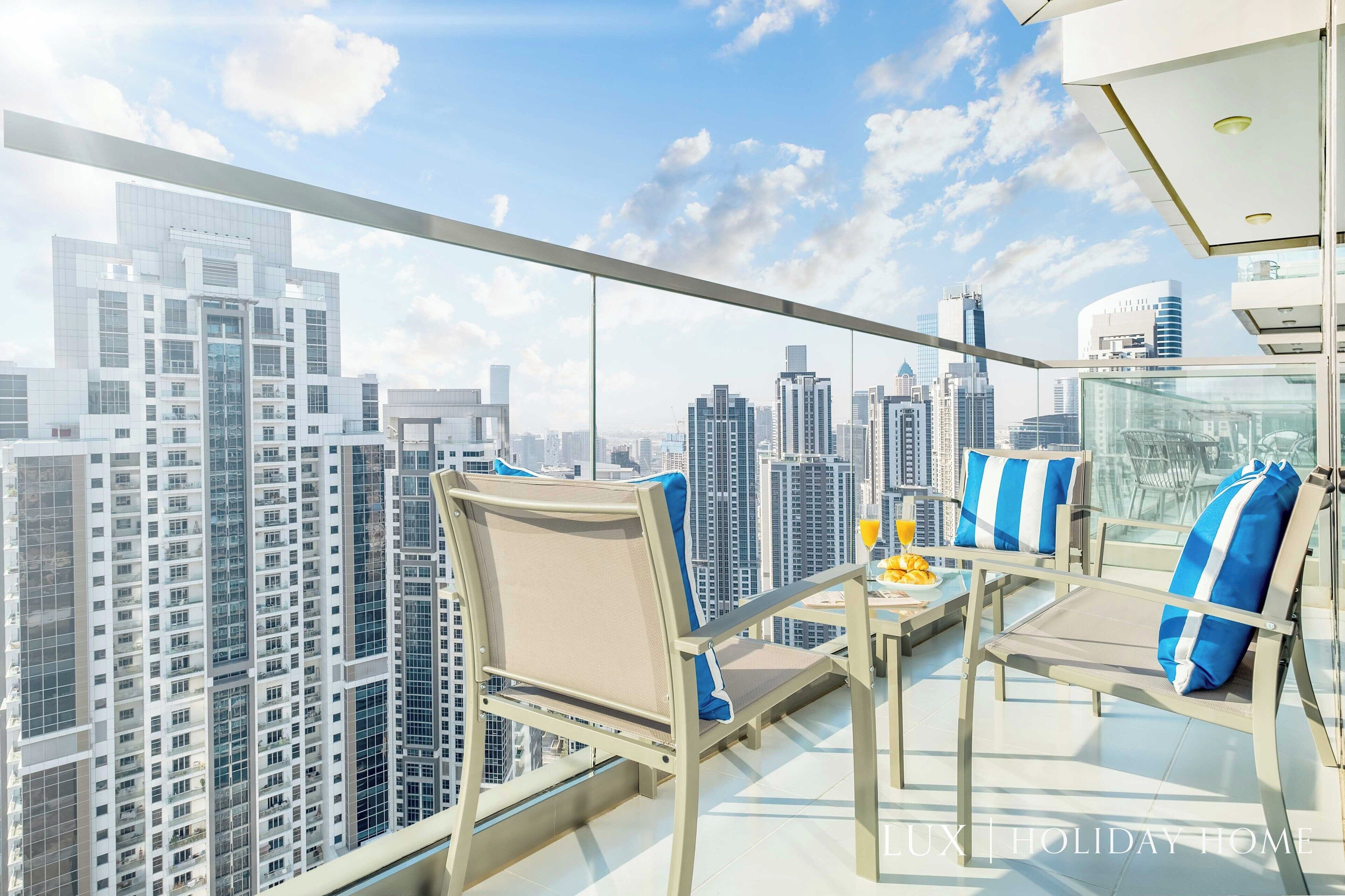 A high-rise balcony with a city view, furnished with two chairs and a table, and a glass railing.