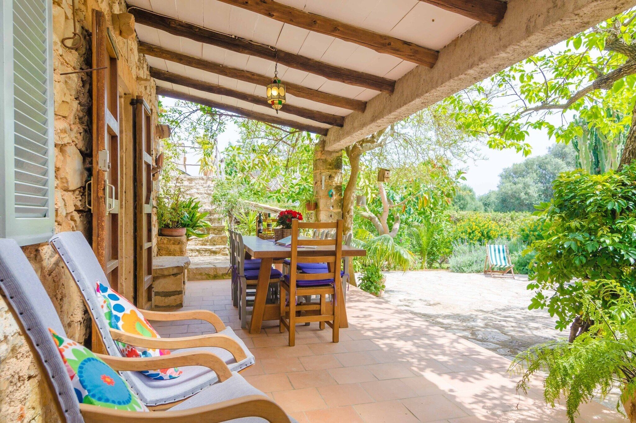An outdoor patio with a wooden table and chairs, surrounded by lush greenery and a stone wall.