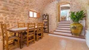 A stone-walled room with a wooden dining table and chairs, a bookshelf, and a staircase leading to another area.
