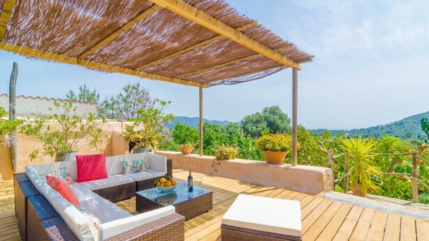 A covered outdoor seating area with wicker furniture and a table, surrounded by potted plants and overlooking a scenic landscape.