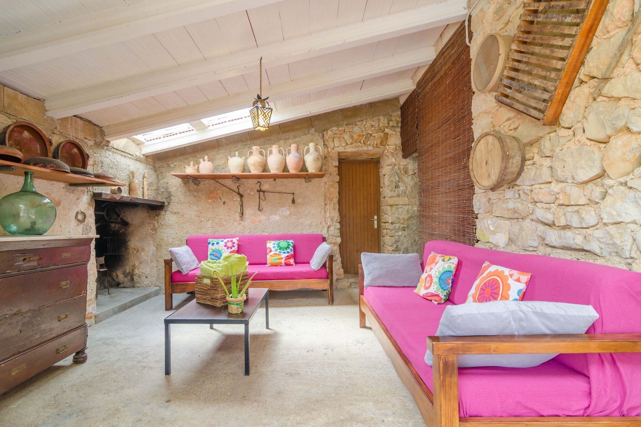 A rustic room with stone walls, a wooden bench, a pink sofa, and a table with a vase and a basket.