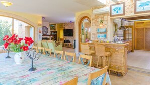 A dining area with a table set for four, surrounded by wicker chairs. A television is mounted on the wall in the background, and there is a stone wall with framed artwork above the counter.