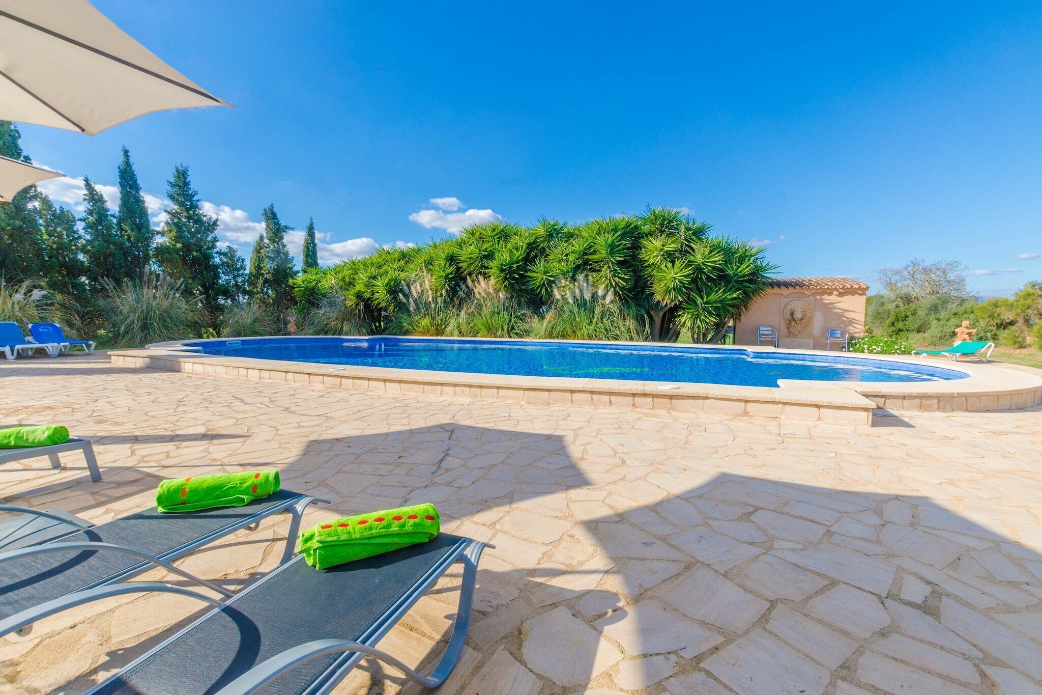 Une piscine entourée de verdure et dotée d’une terrasse en pierre, avec des chaises longues et des parasols.