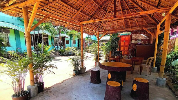 An outdoor covered patio with wooden tables and chairs, surrounded by potted plants and a thatched roof structure.