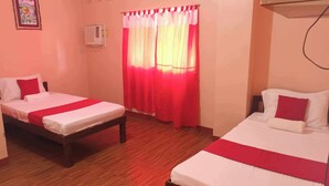 A room with two beds, each with white bedding and red accents, wooden flooring, a wall-mounted air conditioner, and a window with red curtains.