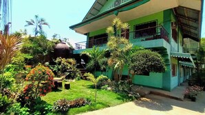 A two-story house with a green facade, a balcony on the upper floor, and a garden with various plants and a large water tank.