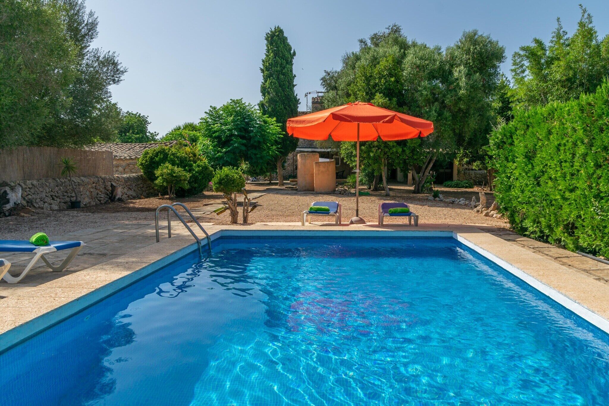 A rectangular swimming pool with clear blue water, surrounded by a sandy area with lounge chairs and an orange umbrella.