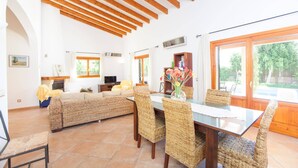 A spacious living area with a dining table, wicker furniture, a sofa, and large windows offering a view of greenery.