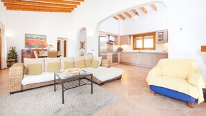 A spacious living area with a sofa, a yellow armchair, and a dining table. A kitchen is visible in the background with wooden cabinets and a window.