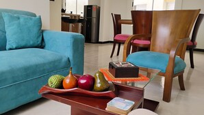 A living room with a blue sofa, wooden chairs, a coffee table with decorative items, and a kitchen in the background.