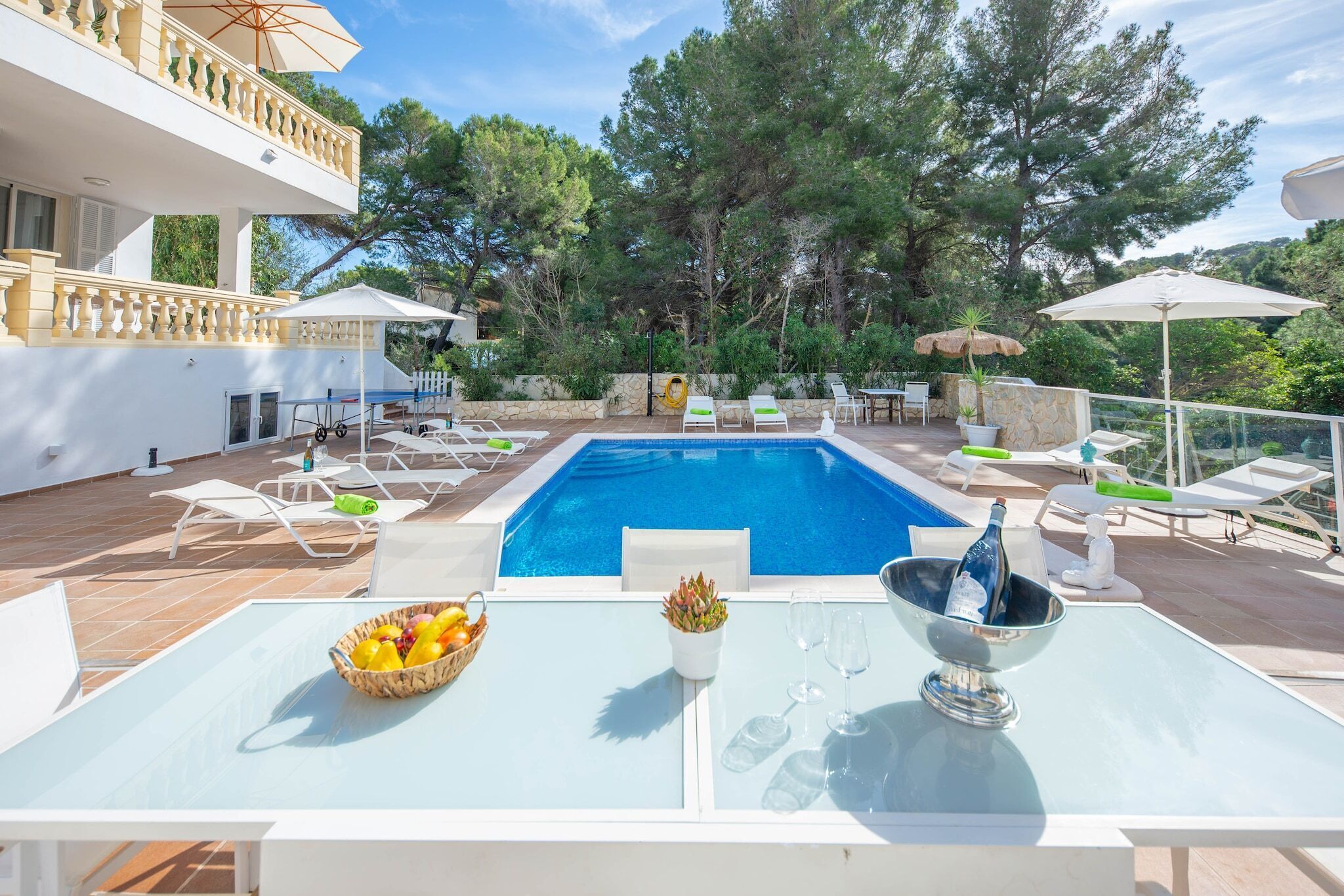 Un área junto a la piscina con una piscina azul cristalina, sillas de descanso y una mesa con un frutero y una botella.