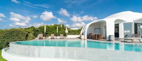 A modern house with a large swimming pool, outdoor seating, and a lush green landscape.