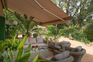 Een overdekt terras met rieten meubels en een tafel, omgeven door weelderige begroeiing en bloempotten.