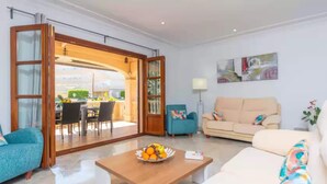 A modern living room with a sofa, a coffee table with fruit, and a dining area visible through an open door.