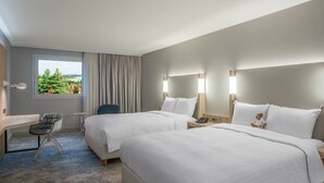 Hotellihuone, jossa on kaksi sänkyä, työpöytä, tuoli ja näkymä vehreään maisemaan.