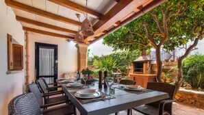 A dining area with a long table set for four, surrounded by wicker chairs. There is a pergola overhead with hanging lanterns, and a tree is visible outside the window.