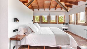 A bedroom with a wooden ceiling, a bed with white bedding, a bedside table, a chair, and a window with a view of greenery.