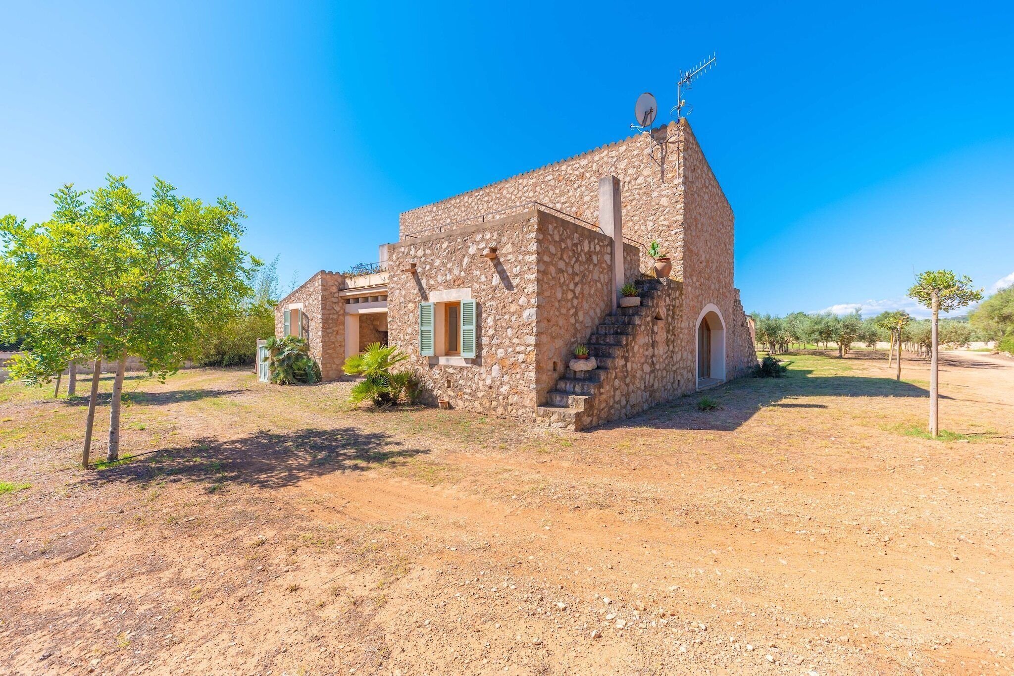 A stone building with a small staircase leading to a door, surrounded by dry grass and a few trees.